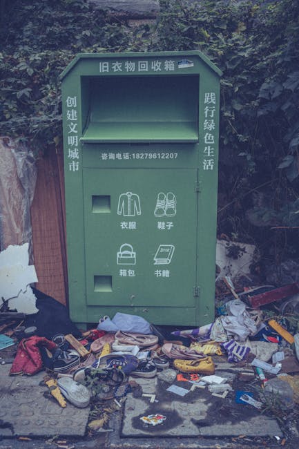 A large pile of black plastic rubbish bags filled with waste is stacked against a tiled wall in an urban environment. Some bags are torn or partially open, revealing plastic bottles and other miscella